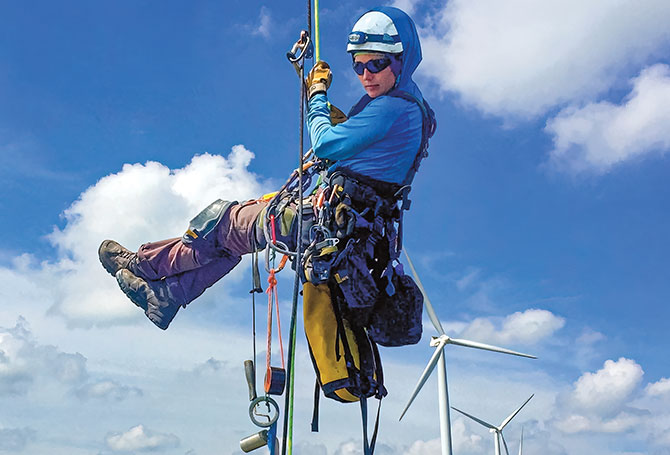 A person in a helmet hanging from ropes and a harness with wind turbines in the background
