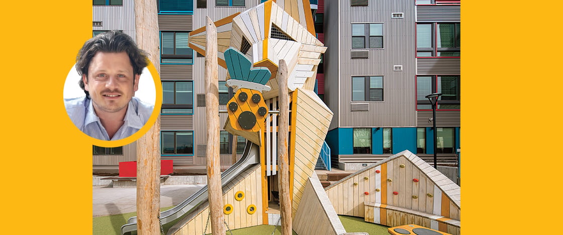 A man's portrait beside a tall wooden playground structure in an urban courtyard
