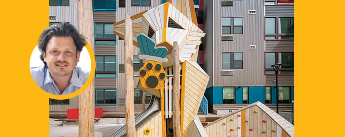 A man poses next to a colorful wooden playground structure in front of a modern apartment building