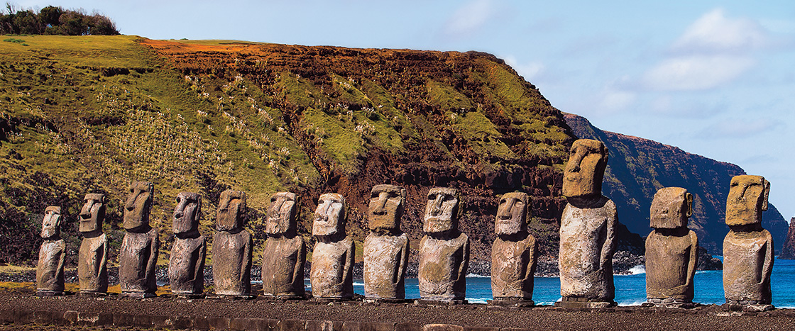 A row of moai stone statues stand along the coast of Easter Island with cliffs behind them