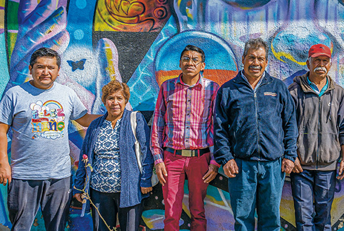 Five people stand in a row in front of a brightly painted mural of blue, purple, & orange shapes