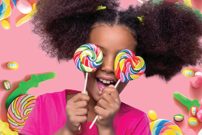 A girl in a pink shirt grins & holds 2 swirled lollipops over her eyes against a pink background