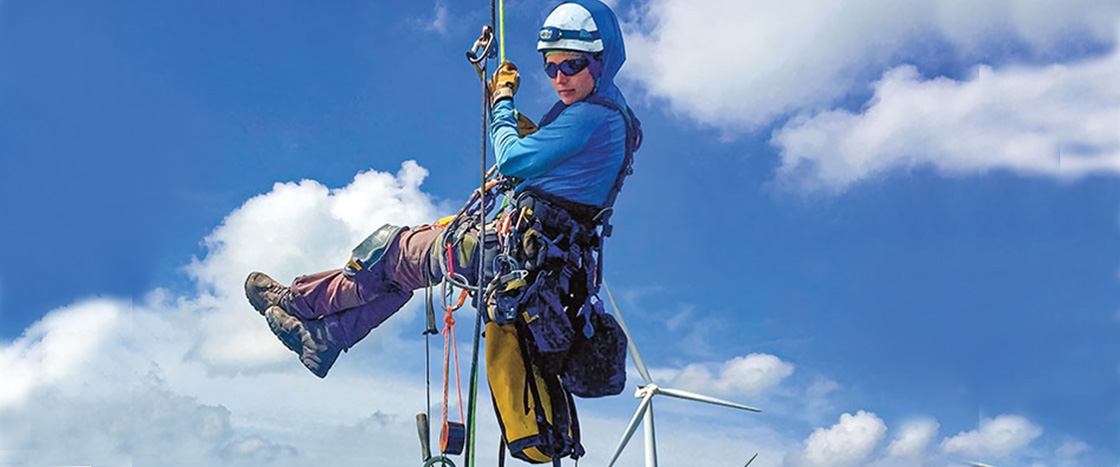 A woman carrying gear and wearing a harness is being lifted towards some turbines